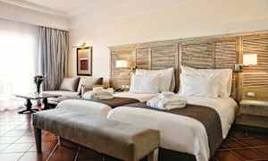 Et hotelværelse med to separate senge, en sofa, et bord med en blomster vase og et vindue med gardiner. (AI-genereret tekst)