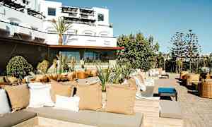 Et udendørs loungeområde med siddegrupper, puder og planter, med en moderne bygning i baggrunden. (AI-genereret tekst)