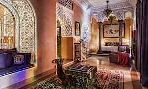 Et elegant indrettet rum med dekorative gipsmønstre, arkitektoniske buer, orientalske møbler og varm belysning. Et bord formet som en kamel står på et farverigt tæppe. (AI-genereret tekst)