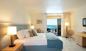 Et hotelværelse med en stor seng, to sengelamper, et skrivebord med en stol og en balkon med udsigt over havet. (AI-genereret tekst)