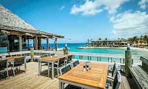 En udendørs terrasse med borde og stole ved en bygning med tag af træplanker. I baggrunden ses havet, en sandstrand og bygninger tæt på vandet. (AI-genereret tekst)