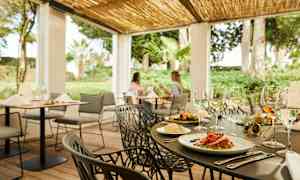 En udendørs restaurant med dækkede borde og grønne planter i baggrunden. (AI-genereret tekst)