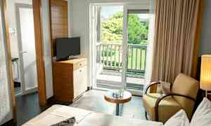 Et hotelværelse med seng, lænestol, bord, TV og en altan med udsigt til en frodig have. (AI-genereret tekst)