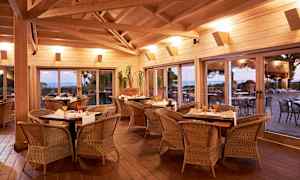 Indendørs miljø med spiseborde og stole af rotting, store vinduer med udsigt til en terrasse og landskab udenfor. (AI-genereret tekst)