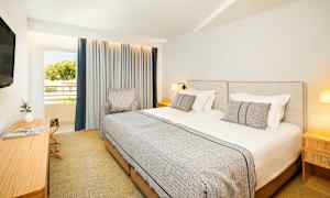 Hotelværelse med to enkeltsenge, en stol, et skrivebord og et TV samt et vindue med gardiner. (AI-genereret tekst)