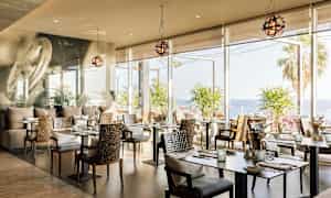 En restaurant med moderne borde og stole, store vinduer med udsigt over havet og palmer. (AI-genereret tekst)