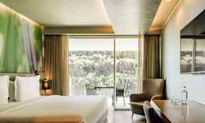 Et hotelværelse med en dobbeltseng, siddegruppe, et bord med stole samt en stor glasdør, der åbner op til en balkon med udsigt over natur og klipper. (AI-genereret tekst)