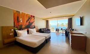 Hotelværelse med to enkeltsenge, sofa, bord og stole. Vindue med udsigt til palmer og hav. (AI-genereret tekst)