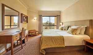 Billede af et hotelværelse med en dobbeltseng, skrivebord og stole samt en altan med udsigt. (AI-genereret tekst)