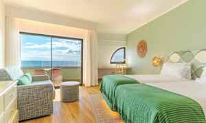 Interiør af et hotelværelse med grønne detaljer, to senge, en siddegruppe og en altan med havudsigt. (AI-genereret tekst)