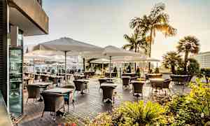 Udendørsterrasse med borde og stole under hvide parasoller, omgivet af grønne planter og palmer. (AI-genereret tekst)