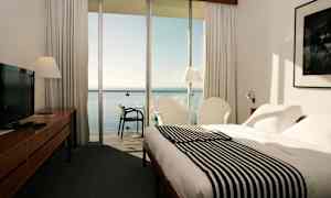 Hotelværelse med seng, lænestole og udsigt over havet gennem store glasvinduer. (AI-genereret tekst)