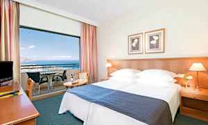 Hotelværelse med dobbeltseng, natbord og balkon med udsigt over havet. (AI-genereret tekst)