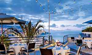 Udendørs spisested med dækkede borde og stole, omgivet af lyskæder og med udsigt over byen og havet. (AI-genereret tekst)