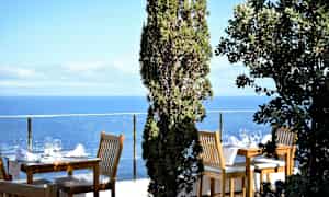Haveudsigt med havet og opsatte borde med stole omgivet af grønne planter. (AI-genereret tekst)