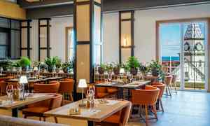 En velbelyst restaurantinteriør med borde, stole og grøn planteudsmykning. I baggrunden er der store vinduer, der viser udsigt over en bygning med et klokketårn. (AI-genereret tekst)