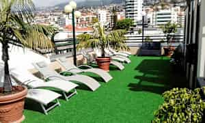 En terrasse med kunstgræs, liggestole og potteplanter. I baggrunden ses byens bygninger. (AI-genereret tekst)