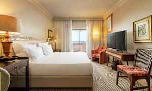 Et hotelværelse med en stor seng, natbord, sofa, TV og et skrivebord. Gardiner og glasdøre fører til en balkon. (AI-genereret tekst)