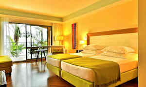 Et hotelværelse med to enkeltsenge, grønne tæpper og en siddegruppe, med udsigt til udendørs palmer gennem store glasvinduer. (AI-genereret tekst)