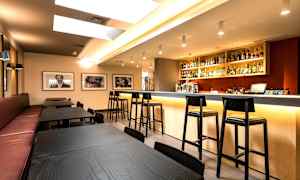 Et moderne barområde med primært sorte borde og stole samt en godt oplyst bardisk fyldt med flasker og glas. (AI-genereret tekst)