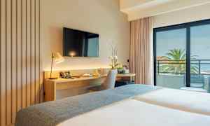 Et hotelværelse med skrivebord, stol og TV, samt en balkon med udsigt over palmer og havet. (AI-genereret tekst)