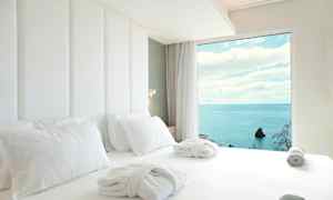 Hotelseng med hvide lagner og badekåber, med havudsigt gennem et vindue. (AI-genereret tekst)