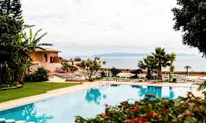 Billede af en udendørs pool omgivet af grønne områder, med liggestole og en bygning i baggrunden. (AI-genereret tekst)