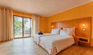 Et hotelværelse med to senge beklædt med hvidt sengetøj, et hovedgærde i træ og en balkon med udsigt. Værelset har gule vægge og trægulv. (AI-genereret tekst)