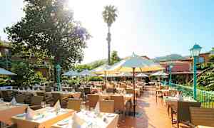 En udendørs terrasse med opdækkede borde omgivet af parasoller og grønne planter. I baggrunden ses en bygning omgivet af træer. (AI-genereret tekst)
