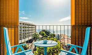 To blå stole og et bord på en balkon med udsigt over havet og omkringliggende bygninger. (AI-genereret tekst)