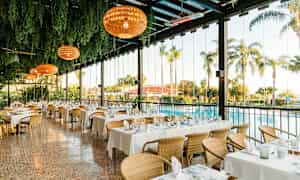 En lys restaurant med opdækkede borde, omgivet af grønne omgivelser og udsigt til en pool og palmer. (AI-genereret tekst)