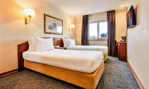 Billede af et hotelværelse med to enkeltsenge, et vindue med gardiner, en vægdekoration og et skrivebord med en stol. (AI-genereret tekst)