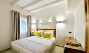 Et lyst soveværelse med en dobbeltseng dekoreret med to gule puder og et sengetæppe. Vinduerne har gardiner, og der er en arbejdsstation med skrivebord og lampe. (AI-genereret tekst)