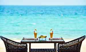 To rottingstole omkring et bord med to glas juice og en skål med frisk frugt på en tropisk strand. (AI-genereret tekst)