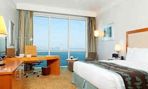 Et moderne hotelværelse med en stor seng, skrivebord og udsigt gennem store vinduer mod havet og en by i baggrunden. (AI-genereret tekst)