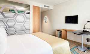 Et hotelværelse med en seng dækket i hvide og beige farver. Værelset indeholder også et skrivebord, et fjernsyn på væggen og et minikøleskab. (AI-genereret tekst)