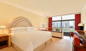 Hotelværelse med dobbeltseng, natborde og udsigt gennem store vinduer over en byens skyline. (AI-genereret tekst)