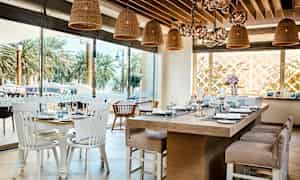 Indretning af en restaurant med borde dækket med porcelæn og glas samt rottinglys hængende fra loftet. Udenfor ses palmer. (AI-genereret tekst)