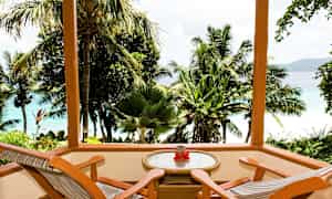 Træterrasse med to stole og et bord med udsigt over tropiske palmer og blåt hav. (AI-genereret tekst)