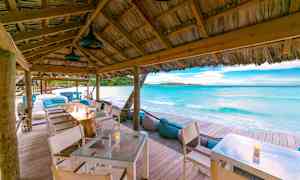 Et strandnært udendørsområde med borde under et trætækt tag nær klart, blåt hav og sand. (AI-genereret tekst)