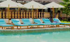 Billede af en swimmingpool med flere blå liggestole og parasoller i forgrunden og en bygning med træbalustrade i baggrunden. (AI-genereret tekst)