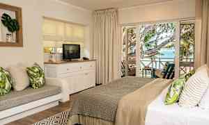 Billede af et hotelværelse med en komfortabel dobbeltseng og et siddeområde, et TV på et hvidt møbel og udsigt over havet fra en møbleret altan. (AI-genereret tekst)