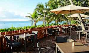 Udendørs serveringsområde ved havet med borde, stole og parasoller, omgivet af frodig bevoksning og palmetræer. (AI-genereret tekst)