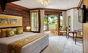 Soveværelse med dobbeltseng, dekoreret med farverige tekstiler og vægkunst, har træpanelvægge og et stort vindue med udsigt over en frodig veranda. (AI-genereret tekst)