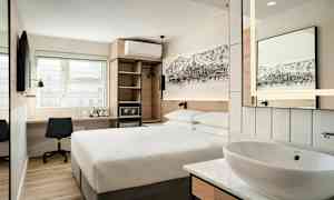 Et moderne hotelværelse med en dobbeltseng, et skrivebord med stol, et vægmonteret TV, samt en åben garderobe og en lille håndvask med spejl. (AI-genereret tekst)