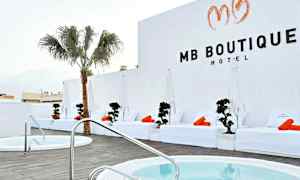 Et hotelområde med hvide liggestole og orange håndklæder ved en pool, samt en palme og hvide vægge. (AI-genereret tekst)