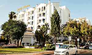 Hvid bygning med hotelskilt, omgivet af grønne områder og parkerede biler. (AI-genereret tekst)