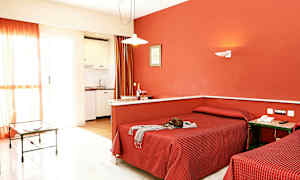 Et hotelværelse med to enkeltsenge, røde sengelinned og en rød væg, samt et lille køkken og et glasbord i baggrunden. (AI-genereret tekst)