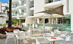 Et udendørsområde med borde, stole og parasoller foran en bygning. (AI-genereret tekst)