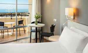 Et hotelværelse med en seng, bord og stole tæt på en altan med havudsigt. (AI-genereret tekst)